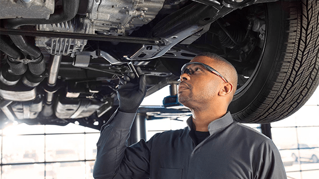 Mechanic inspecting car undercarriage, wearing safety goggles and gloves in a workshop.