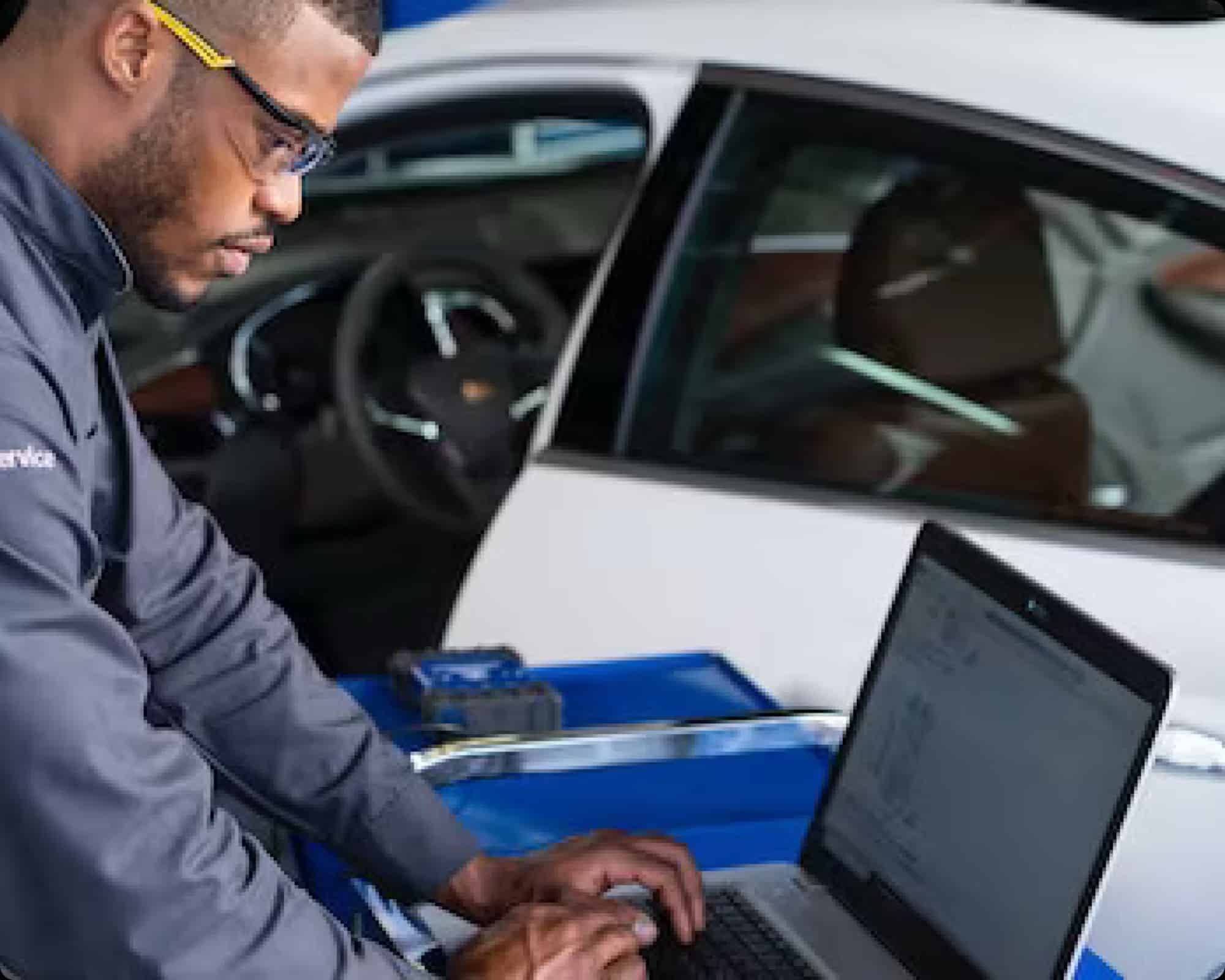 Mechanic using a laptop for diagnostics beside a white car in an auto repair shop.
