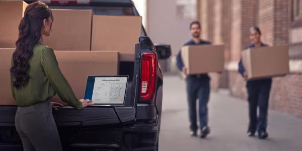 Woman using a laptop on a truck tailgate while two workers carry boxes in an alleyway.