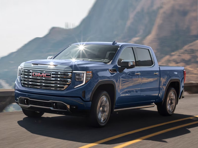 Blue GMC Denali truck driving on a scenic mountain road, reflecting sunlight.