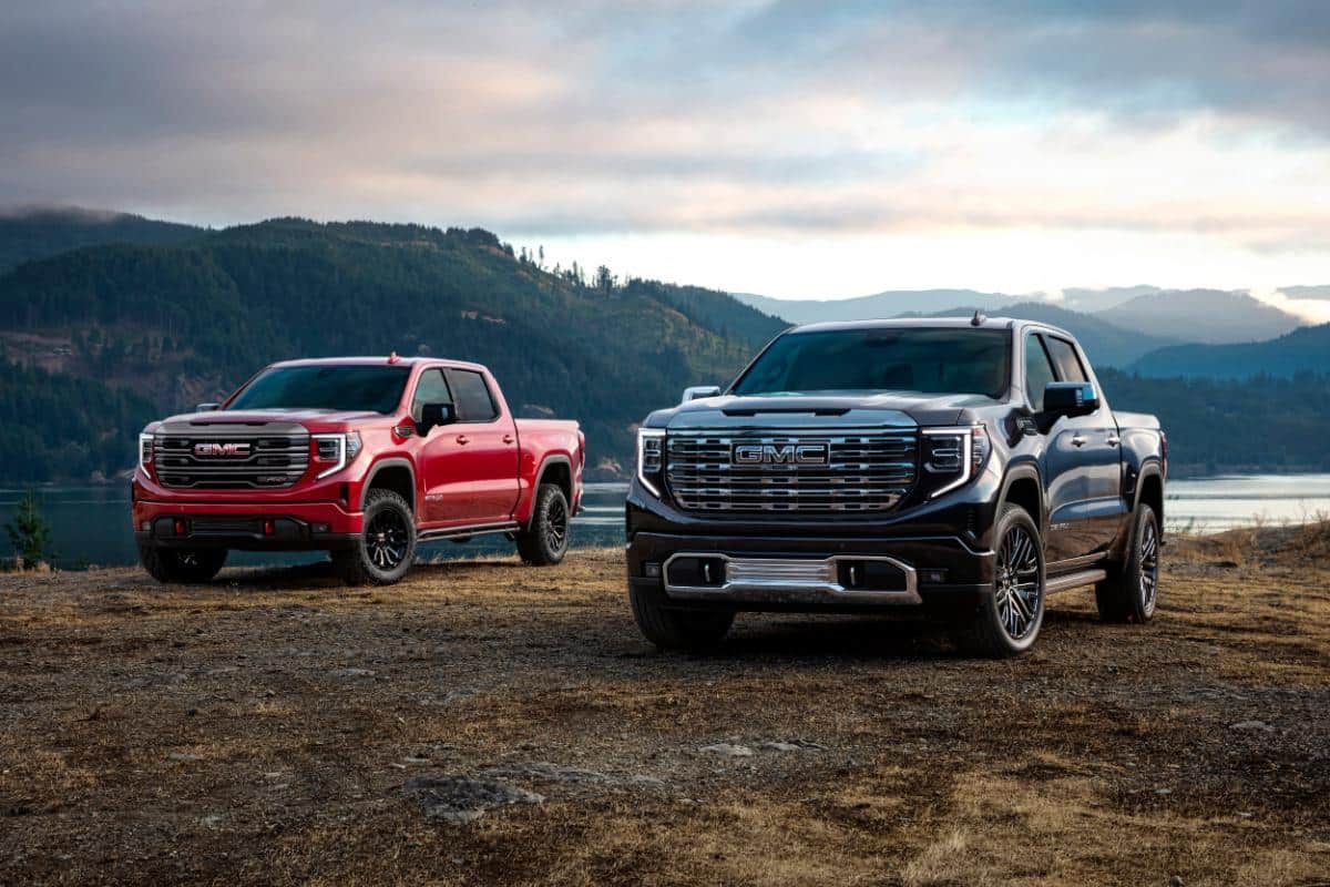 Two GMC trucks parked on a scenic landscape with mountains and a lake in the background.