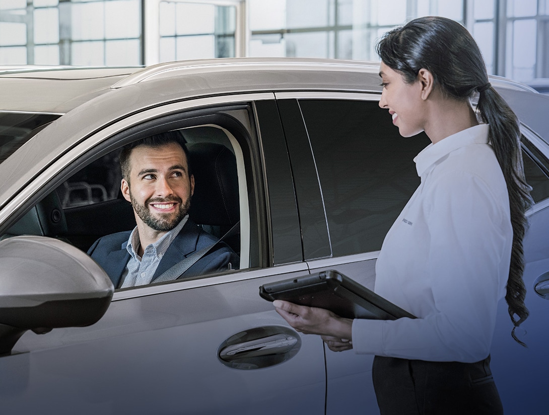 Man in car interacting with a smiling woman holding a tablet, in an automotive service setting.