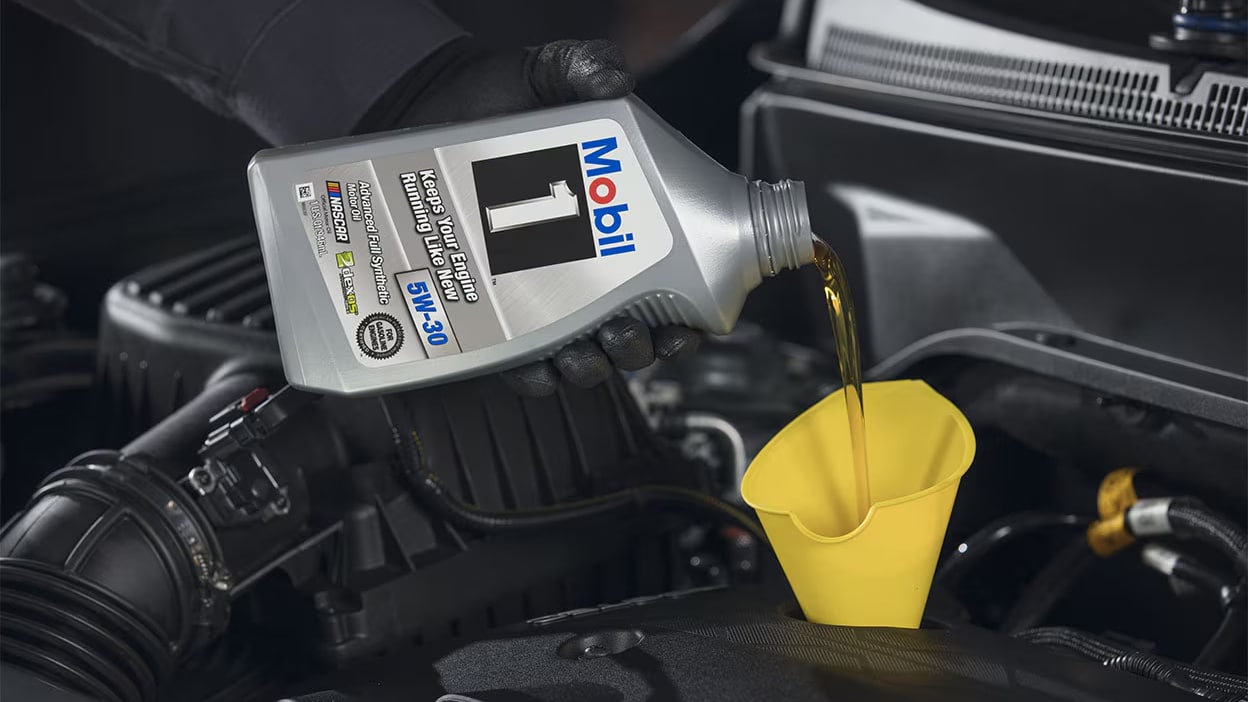 Pouring Mobil 1 synthetic oil into a car engine using a yellow funnel.