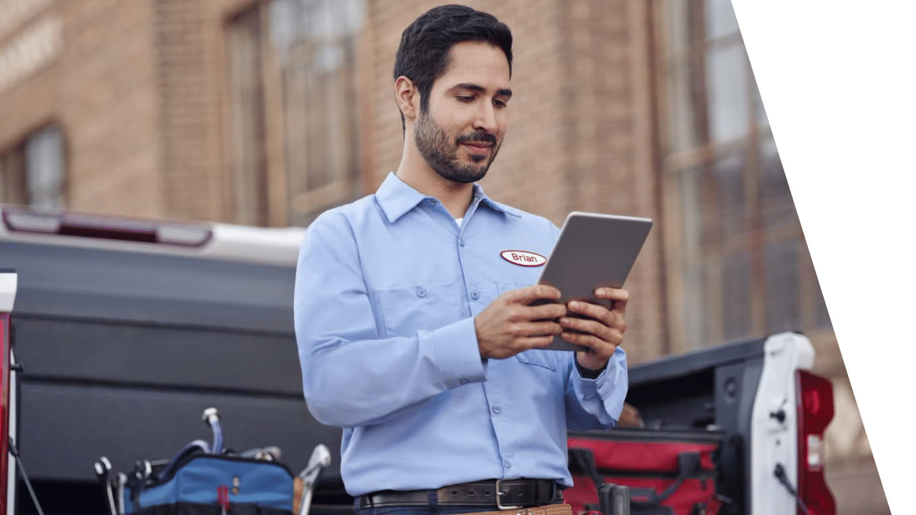 A technician in a blue shirt uses a tablet near a truck with tools outside.