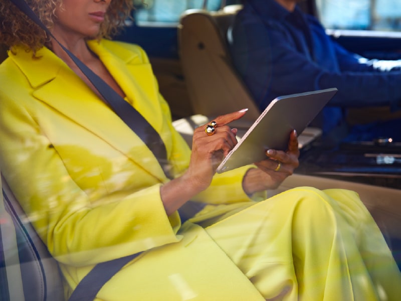 Woman in yellow suit using tablet in a car, seated next to a driver.