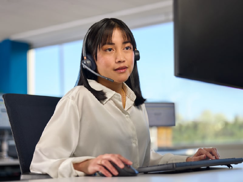 A woman in a white shirt wearing a headset, working on a computer in an office.