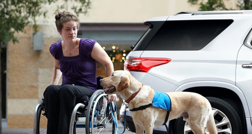 A woman in a wheelchair with a service dog in a blue vest beside her, parked SUV in the background.