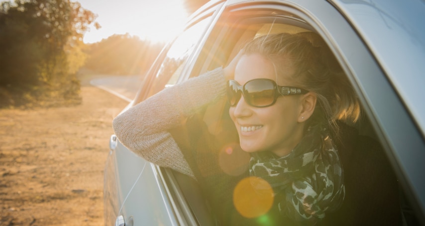 Woman with sunglasses smiling and leaning out of a car window on a sunny day.
