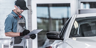 Mechanic inspecting a vehicle at a garage, wearing a cap and overalls with a clipboard in hand.