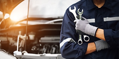 Mechanic in uniform holding wrenches stands by car with open hood, sunlight in background.