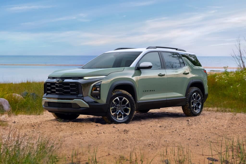 Green SUV parked on a sandy beach with grassy surroundings and a serene sea backdrop.