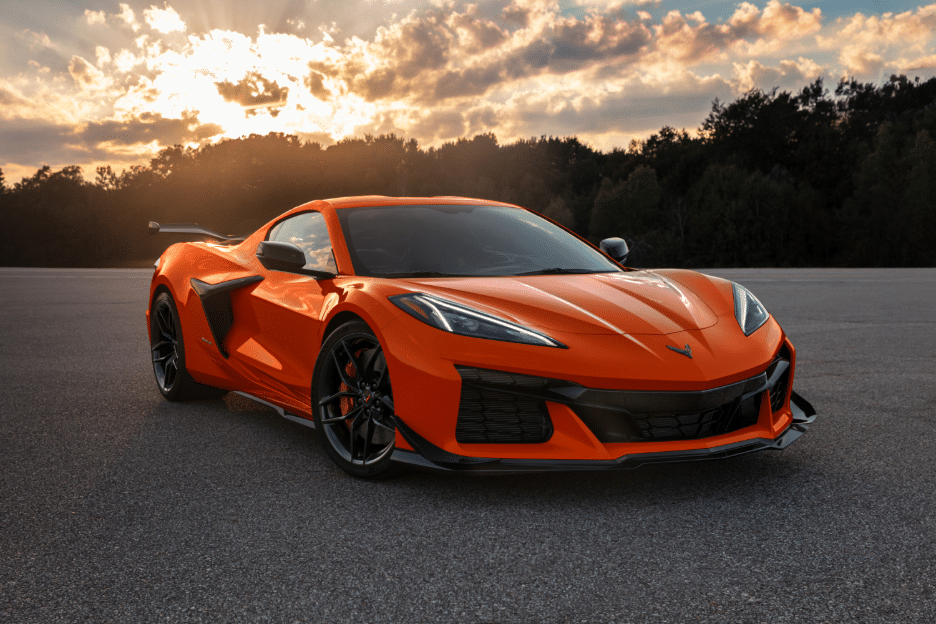 Orange sports car parked on asphalt with dramatic sunset and trees in the background.