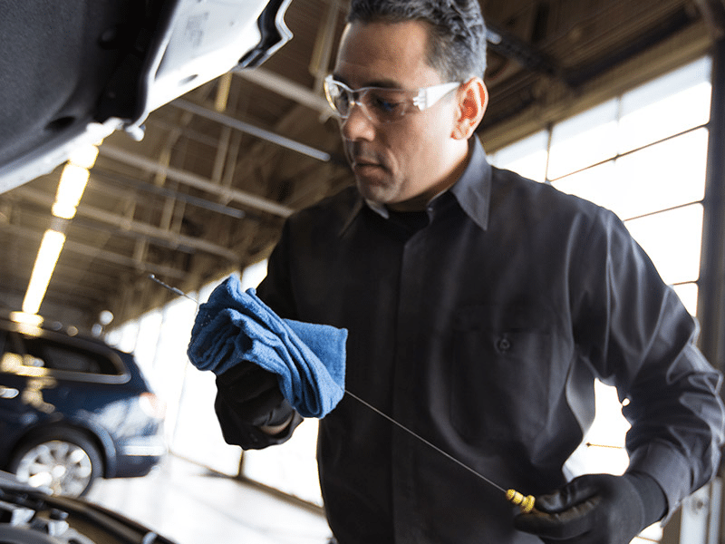 Mechanic checking car oil with dipstick in a service center, wearing safety glasses and a "Certified Service" shirt.
